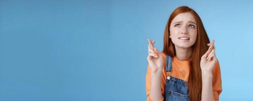 Worried unconfident young redhead intense cute girl frowning nervously waiting improtant moment cross fingers good luck bite lip anxiously frowning look upper left corner praying wish come true photo