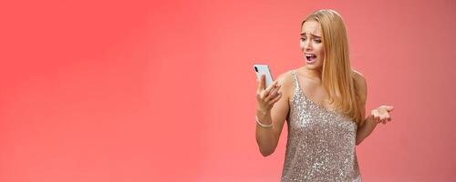 Troubled concerned arrogant young blond woman complaining yelling smartphone cannot call friend no signal holding smartphone look mobile display pissed moody arguging, red background photo