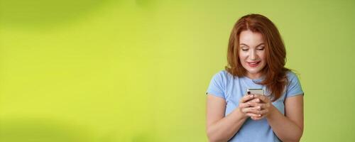 Intrigued excited tempting redhead middle-aged woman shopping online smartphone hold mobile phone look pleased entertained gadget screen smiling delighted playing game found perfect app photo