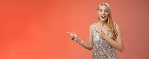 Excited carefree happy young attractive woman joyful attend awesome party in silver beautiful glittering dress pointing left suggesting come together inviting dance smiling cute red background photo