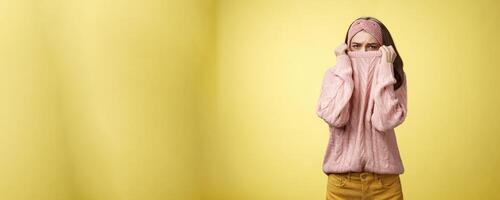 Sad silly, gloomy cute young girl hiding face in sweater collar pulling clothes on nose frowning displeased, unhappy, looking indignant and reluctant standing disappointed over yellow background photo