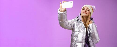 Happy delighted carefree fair-haired charming european woman in silver winter jacket hat raising smartphone horizontally taking selfie smiling mobile phone display, purple background photo
