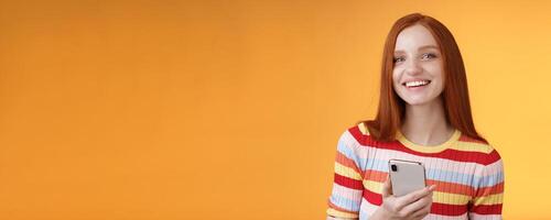 Friendly enthusiastic young redhead girl blue eyes using smartphone turn camera answer smiling broadly telling who sent message standing delighted orange background messaging, texting boyfriend photo