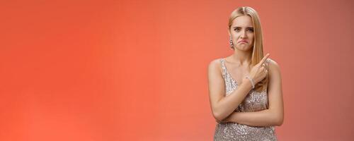 Jealous displeased angry young revengeful blond ex-girlfriend in luxurious silver shiny dress frowning pouting pointing upper right corner displeased pissed standing fed up upset red background photo