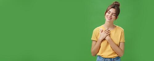 Happy kind and timid young attractive woman with gapped teeth smiling sincere and grateful closing eyes pressing hands on chest in thankful pose, daydreaming, recalling nice tender memories photo