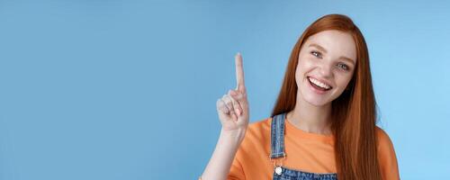 Lively active energized happy smiling hepful redhead female coworker showing you awesome promo pointing index finger up grinning introduce cool product recommend try out blue background photo