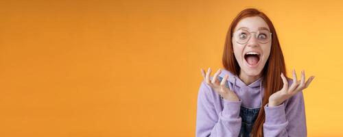 Shocked excited overwhelmed young screaming happy redhead girl wearing glasses winning hear excellent news yelling out loud rejoicing unbelievable luck raising hands spread stare surprised photo
