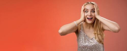 Amused surprised excited young blond attractive woman in silver glittering stylish dress hold head hands widen eyes speechless look camera unbelievable thrill fan see famous person, red background photo
