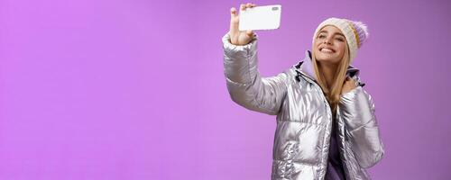 Happy delighted carefree fair-haired charming european woman in silver winter jacket hat raising smartphone horizontally taking selfie smiling mobile phone display, purple background photo