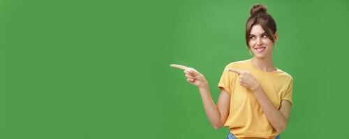 Waist-up shot of charismatic happy and carefree charming woman in yellow t-shirt pointing and looking left enthusiastic and pleased smiling cheerfully posing against green background delighted photo