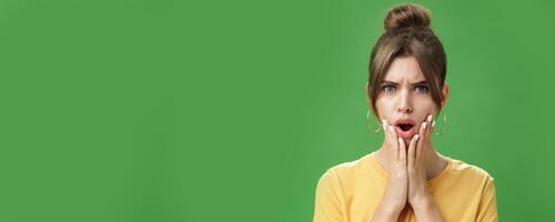Shocked woman reacting to pimple looking disturbed and displeased in mirror holding hands pressed to cheeks open mouth and frowning being disappointed and upset over green background photo