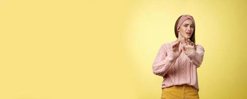 Omg no. Cute young european schoolgirl step back stooping grimacing disgusted and displeased turning away reluctant expressing aversion extending palm in rejection, refusal gesture over yellow wall photo