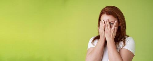 Not peeking. Playful charismatic middle-aged ginger redhead woman close face palms look through fingers one eye stare camera intrigued waiting surprise gift stand green background photo