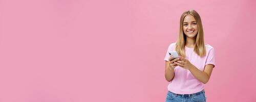 Woman calling taxy via smartphone asking friend address smiling cheerfully at camera holding phone with both hands over chest getting in touch with clients via messages standing over pink wall photo