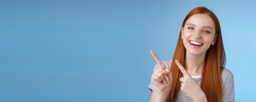Happy carefree smiling redhead caucasian girl ginger straight hairstyle pointing upper left corner laughing joyfully amused cool promo giving recommendation try new product, standing blue background photo