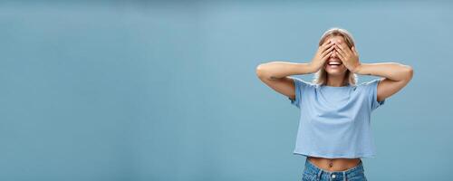 Hide if you can. Portrait of carefree and happy good-looking playful girl with blonde hair smiling broadly while counting ten and playing hide-and-seek with closed eyes over palms near blue wall photo