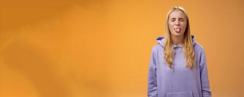 Playful amused charismatic young funny blond girl having fun close eyes showing tongue rebellious fool around unbothered enjoying carefree sunny spring days standing orange background photo