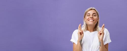 Waist-up shot of hopeful optimistic attractive stylish woman in white t-shirt with tattooed arms winking smiling joyfully while crossing fingers for good luck making wish over purple background photo