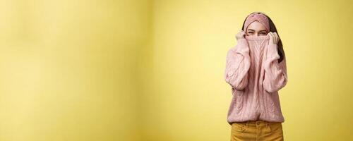 Cute playful young girl hiding in sweater peeking and squinting flirty over top of cloth pulling collar on nose, fooling around having fun happily, being in positive good mood, warming-up on cold day photo