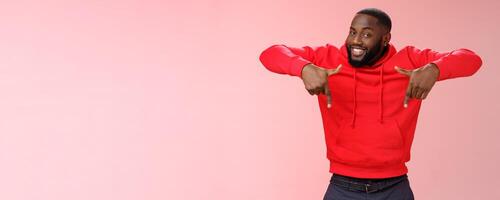 Sassy young charismatic handsome bearded black guy in red hoodie pointing down index fingers smiling broadly showing awesome copy space solution cool product for you, standing pink background photo