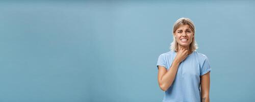 Girl feeling discomfort in throat catching cold or having seasonal allergy touching neck and frowning with clenched teeth and painful expression standing displeased over blue background photo