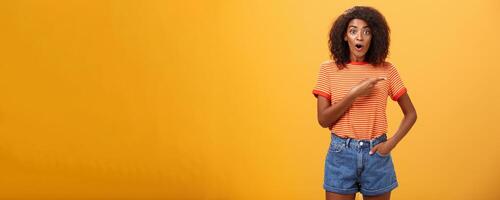 Studio shot of stylish fit female model in denim shorts and t-shirt gasping impressed pointing right while asking question being interested during observing masterclass over orange background photo