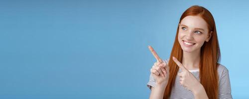 Attractive redhead female model pure clean skin ginger long hair pointing looking left intrigued smiling satisfied indicating interesting advertisement intrigued using new product, blue background photo