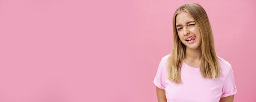 Playful flirty attractive young girl with fair straight hair in t-shirt winking at camera smiling and licking upper lip with tongue feeling enthusiastic and carefree against pink background photo