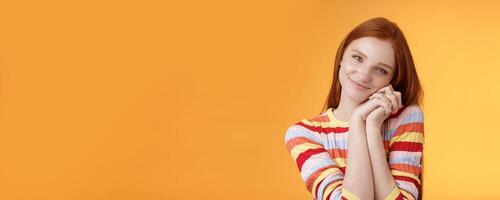 Sweet silly tender redhead young girl leaning palms touched smiling receive charming lovely gift standing thankful look affection sympathy accept dearest romantic gesture, orange background photo