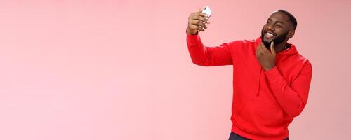 Cheeky cool african american confident bearded guy in red hoodie taking selfie update date app profile photo holding smartphone raised touching face sassy smiling look phone display flirty