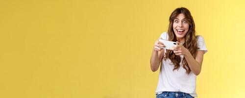 Happy enthusiastic excited cute playful girl winning awesome smartphone game thrilled playing stare camera surprised triumphing smiling happily hold mobile phone horizontal yellow background photo