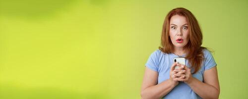 Shocked speechless impressed redhead european middle-aged woman fold lips astonished wow stare camera fascinated react amazed wondered received message hold smartphone intense green background photo