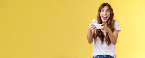 Extremely happy thrilled playful girl gamer playing awesome great new smartphone game hold mobile phone horizontal cheering look camera astonished impressed beating record yellow background photo