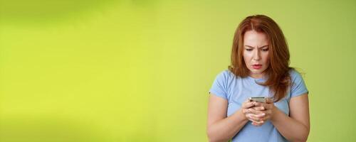 Confused unsure redhead middle-aged woman learn how use social media trying understand emoji look intense focused smartphone display reading important news message stand green background photo
