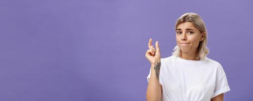 Girl having tiny problem. Concerned attractive blonde girl with tattoo on arm pursing lips in troubled look shaping small or little object, dissatisfied with regret in eyes over purple wall photo