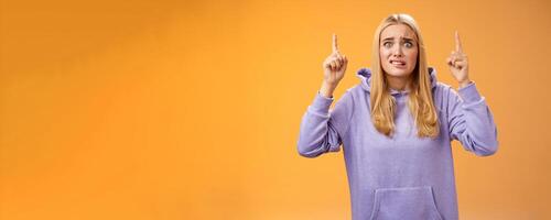 Worried perplexed unsure cute blond woman presenting project standing awkward afraid humiliation crining scared pointing up raised index finger waiting reply nervously, orange background photo
