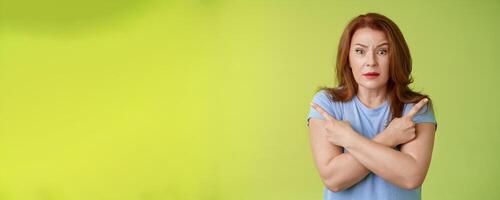 Help me choose. Worried unsure doubtful cute redhead middle-aged silly woman asking advice cross arms body pointing sideways left right look concerned camera make difficult decision green background photo