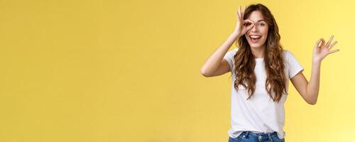 Friendly charismatic enthusiastic happy woman having perfect day show okay ok circle gesture look through ring amused wondered smiling broadly delighted express joy admiration yellow background photo