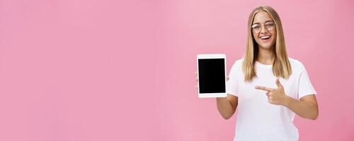 Female freelance programmer proudly showing her app for digital tablet holding gadget pointing at device screen smiling broadly with delighted expression wearing glasses, posing over pink wall photo