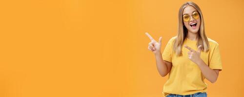 Click her watch my videos. Happy confident and outgoing charming young girl with straight hair in sunglasses and yellow t-shirt pointing at upper left corner promoting product with joyful smile photo