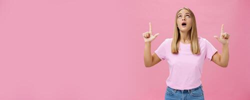 Portrait of amused and impressed curious attractive woman in t-shirt dropping jaw looking and pointing up astonished and intrigued watching interesting object in sky posing against pink background photo
