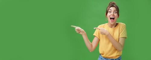 Waist-up shot of enthusiastic and charismatic impressed happy girl laughing and smiling broadly pointing left at funny hilarious thing reacting surprised and entertained, posing over green background photo