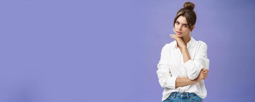 Portrait of tender and feminine stylish woman in white blouse posing sensually and flirty touching chin gazing daring at camera posing against purple background with self-assured expression photo