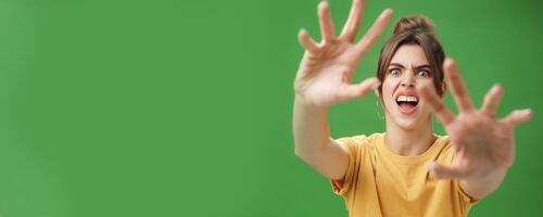 Funny emotive woman making fun face pulling hands forward to attack of grab something frowning opening mouth wanting cover face from camera, grimacing over green background photo