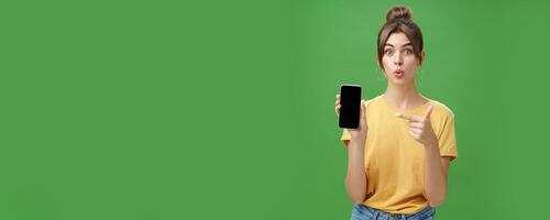 Portrait of intrigued woman cannot wait use new smartphone holding phone and pointing at cellphone screen folding lips with excitement and interest presenting new device model in store over green wall photo