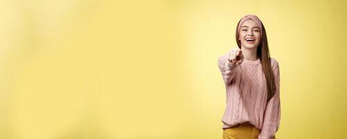 Amused charming tender young european girlfriend mocking having fun of someone pointing finger at camera laughing over funny situation, chuckling, mockering against yellow background photo