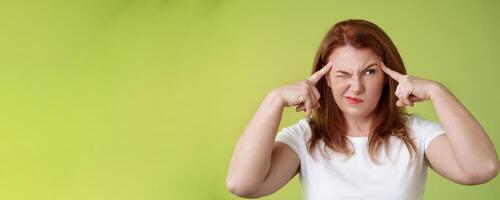 Difficult decision. Perplexed hesitant redhead middle-aged 50s woman trying solve puzzle look puzzled troubled smirk close eyes peek right touch temples thoughtful thinking intense green background photo