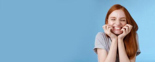 Cheerful carefree giggling ginger girl look happy bright close eyes smiling delighted hold hands cheeks having fun feeling excitement joy triumphing cheering good news, standing blue background photo