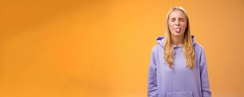 Playful amused charismatic young funny blond girl having fun close eyes showing tongue rebellious fool around unbothered enjoying carefree sunny spring days standing orange background photo