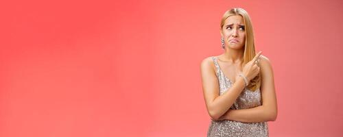 Upset doubtful unsure cute blond funny glamour girl in silver evening dress cringing grimacing hesitant pointing looking upper right corner suspicious, uncertain standing red background photo
